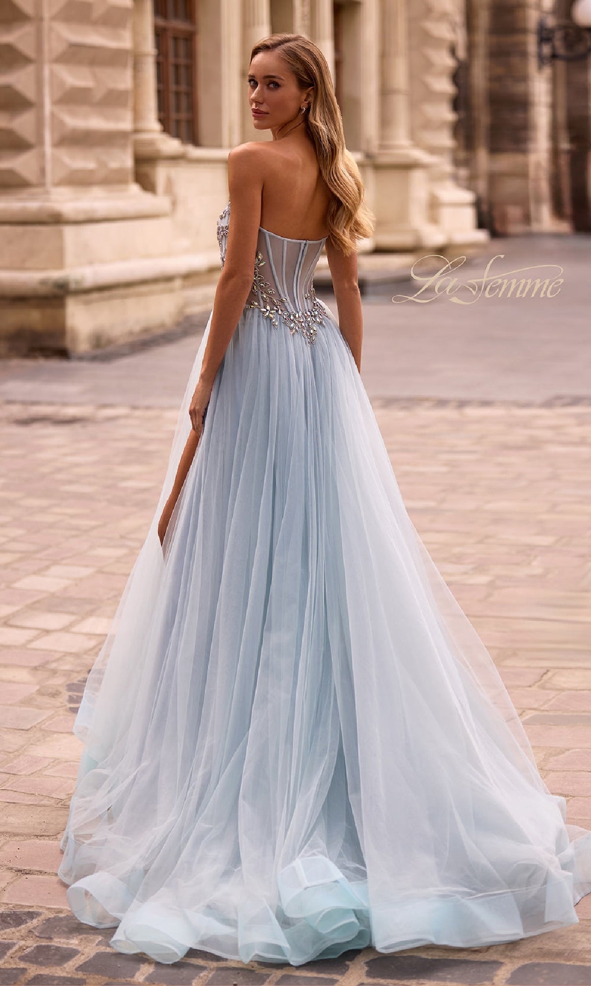 A woman wearing the La Femme 32862 Beaded-Corset Strapless Prom Gown, featuring a beaded corset and light blue tulle A-line skirt, stands on a cobblestone street near ornate buildings.